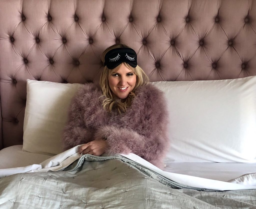 A white woman with MS is in full hair and makeup is sitting in a bed with a pink tufted headboard. She is wearing a pink feather jacket and a sleep mask sits on her forehead. She looks fancy and more high-maintenance than burden.