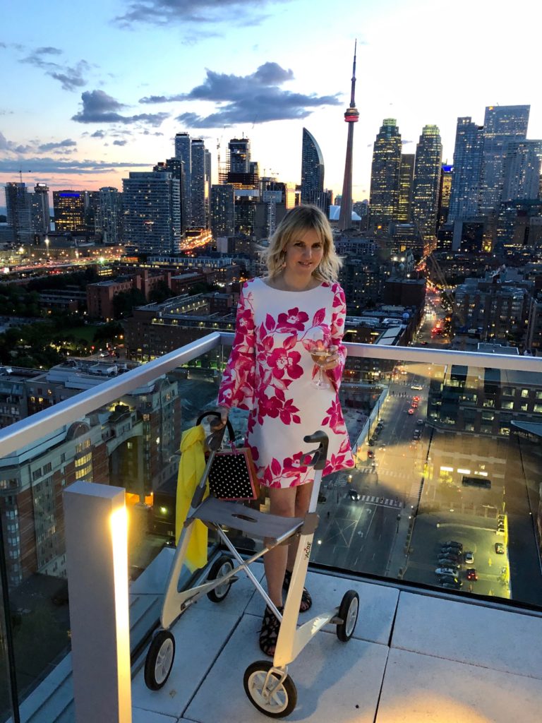 Ardra stands on a rooftop patio with the Toronto city skyline behind her. It is dusk. She looks stylish in a pink and white mini-dress and her oyster-white byAcre rollator.
