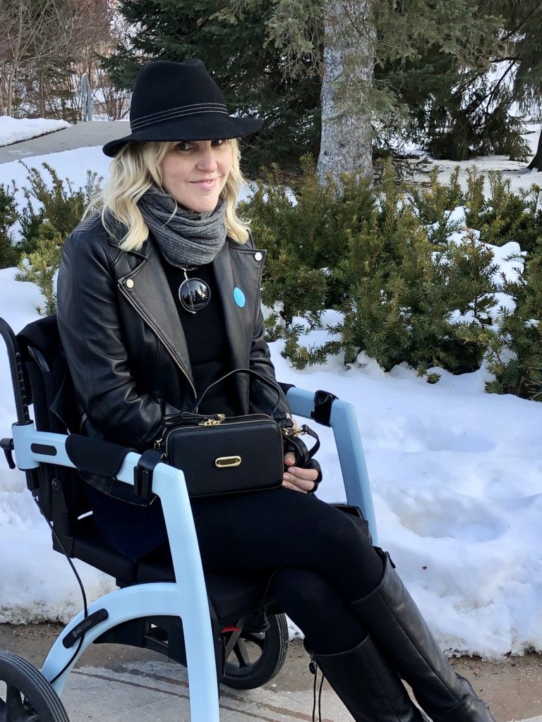 Ardra sits in the Rollz motion in transport chair mode. She is wearing the same leather jacket and a black fedora. She has a black bag from FFORA on her lap.