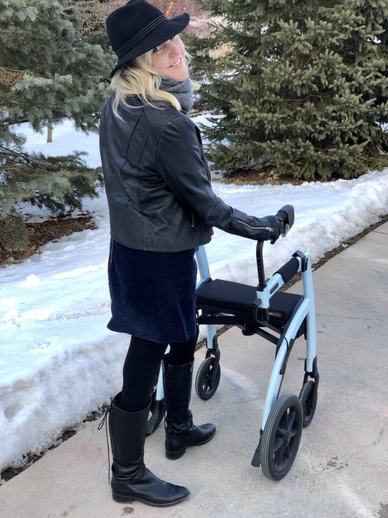 Ardra, a white woman with blonde hair, stands behind her Rollz in rollator mode. She's looking back over her shoulder and smiling. She looks cool in her leather jacket and her confidence with the Rollz Motion  only makes her look cooler.