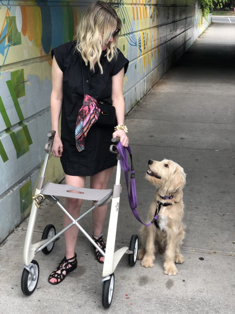 Ardra stands outside, next to some graffiti. She has a white rollator and her dog is looking at her adoringly. She's pretty sure that you can't even bring a service dog to your MRI no matter how much you hate MRIs.