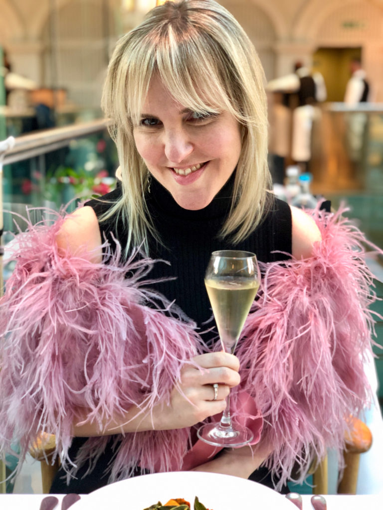 The author, Ardra Shephard, a white woman with shoulder-length blonde hair, is sitting in a glamorous restaurant with waiters in tuxedos in the background. She is wearing a black sleeveless dress and a pink feather boa. She is smiling and holding a glass of champagne and living a fabulous life despite having bowel problems and MS.