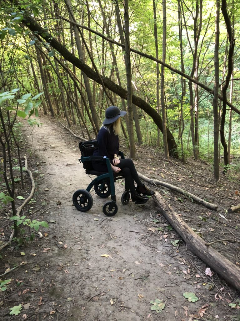 Ardra sits in the Rollz Performance, a dark green, updated version of the Rollz Performance. She is on a trail in the woods and the ground is uneven. She is looking off into the distance, enjoying some time in nature.