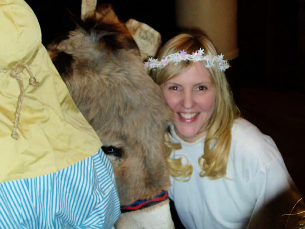A blonde woman in an angel costume is kneeling next to a real donkey in a church Christmas pageant.