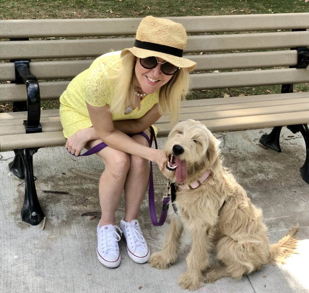 A white woman in a yellow dress sits on a bench next to her golden-doodle puppy. An example of joy during difficult times. You don't have to wait for the pressure to have a happy new year to be happy.