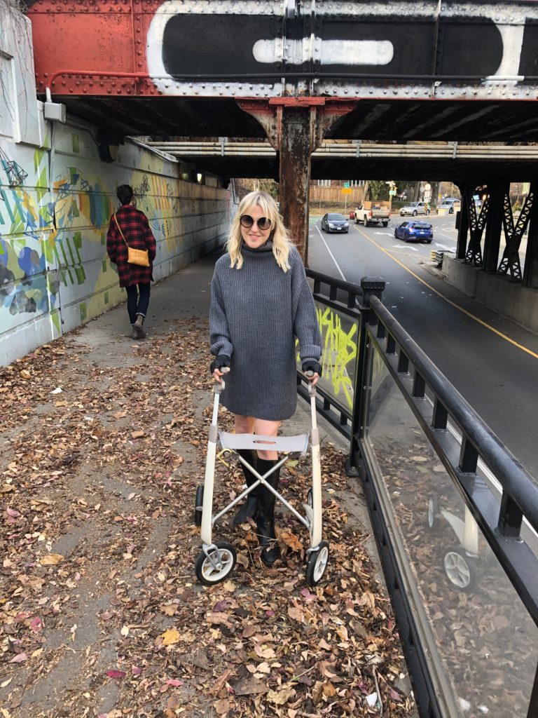 A white woman wearing a grey sweater dress and knee-high boots stands on a sidewalk covered in leaves. She is using a stylish rollator from byAcre. She is smiling.