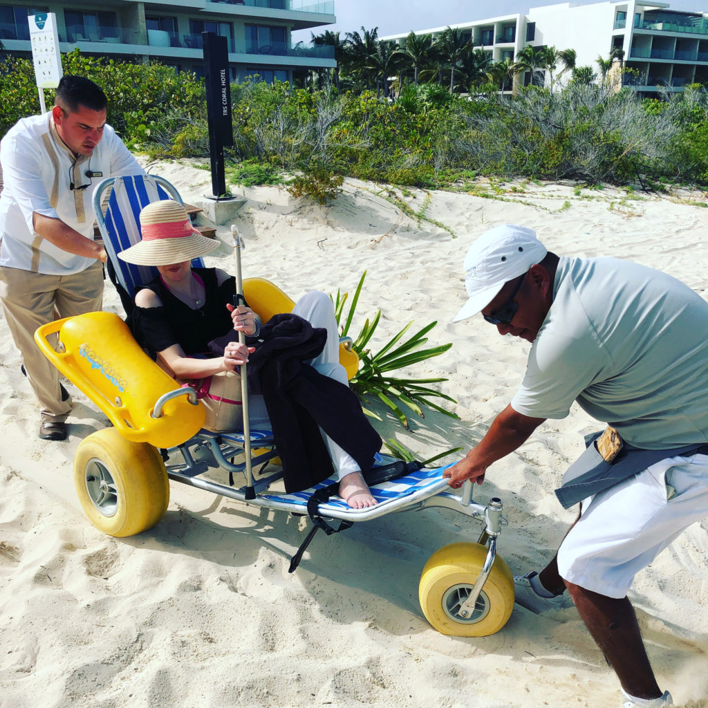 A woman with MS is in a beach wheelchair. She looks fancy. She is being helped by two men. The men are making it look like hard work, but she insists she isn't a burden.