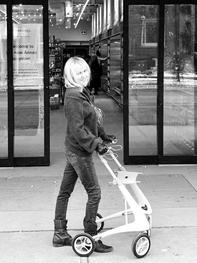 A white woman with MS stands in profile in front of her rollator. She's wearing skinny jeans which truthfully can sometimes be a bit of a burden to get off if you have MS.